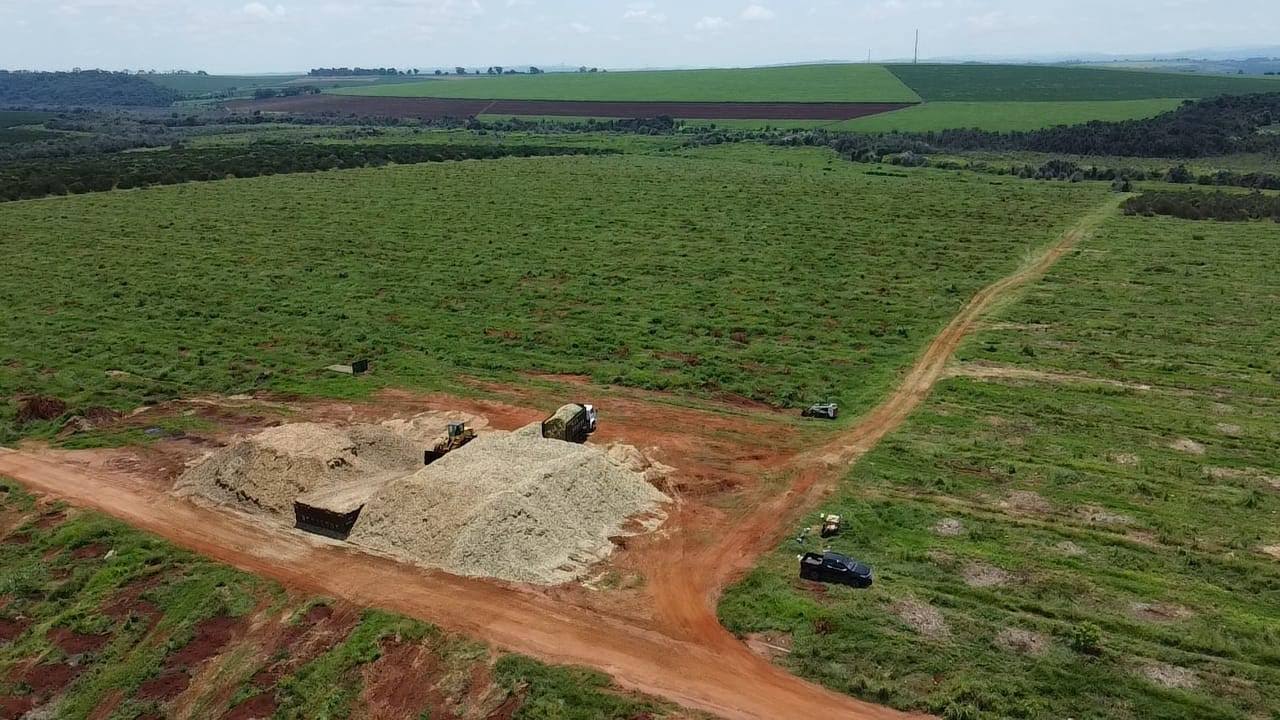Vista aérea de grandes pilhas de cavaco de madeira em área rural, com máquinas e caminhão utilizados no processamento e transporte de biomassa.