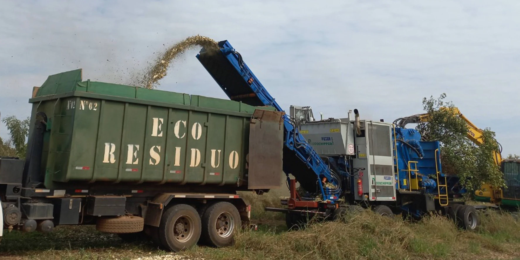 Máquina trituradora processando resíduos de madeira e carregando cavaco de biomassa em caminhão da EcoResíduos durante operação de erradicação de plantação.